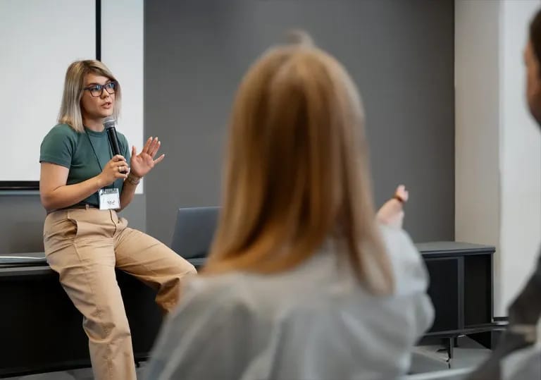 Una relatrice rilassata parla al microfono seduta sul bordo di un tavolo, dimostrando come trasformare la paura di parlare in pubblico in una vera competenza comunicativa.
